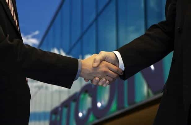 Two people shaking hands as they secure a new deal in front of a building with a large Bitcoin sign on its front.