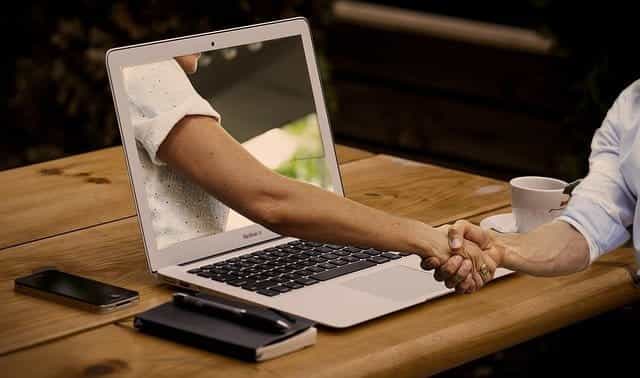 Someone shaking the hand of another person whose arm is coming out of the screen of a laptop, placed on a table and flanked by a smartphone, notebook and coffee mug.