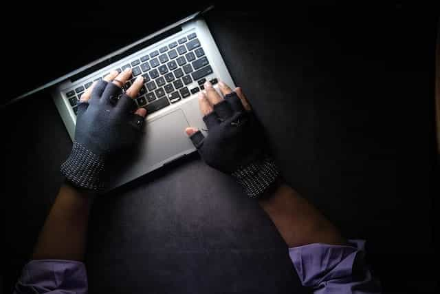 Hands with black finger gloves on them hover over a laptop keyboard in a dark setting.