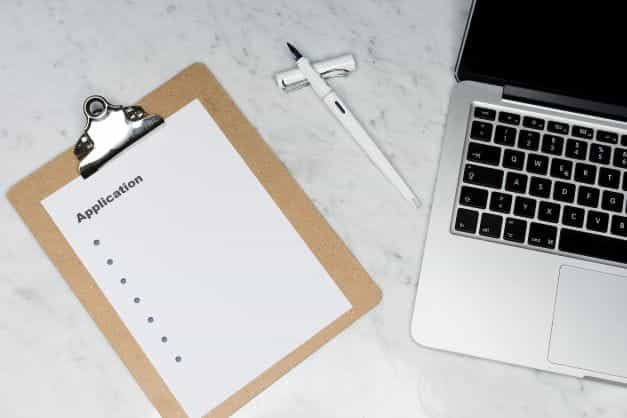 A white paper on a clipboard with the word application as the title, placed next to a laptop.