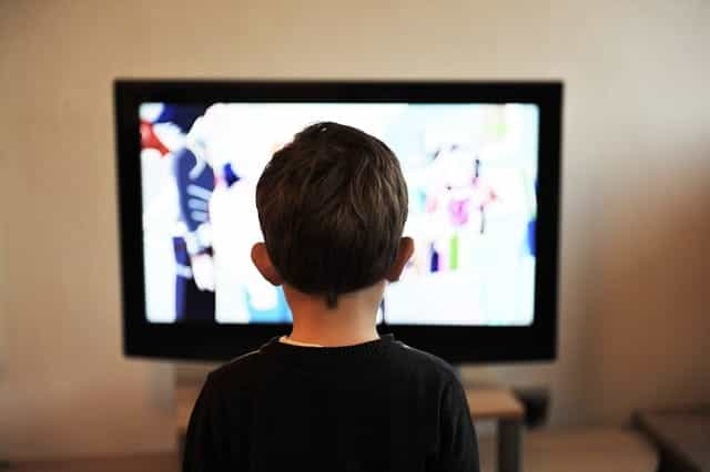 The back of a child sitting in front of and watching a television screen.