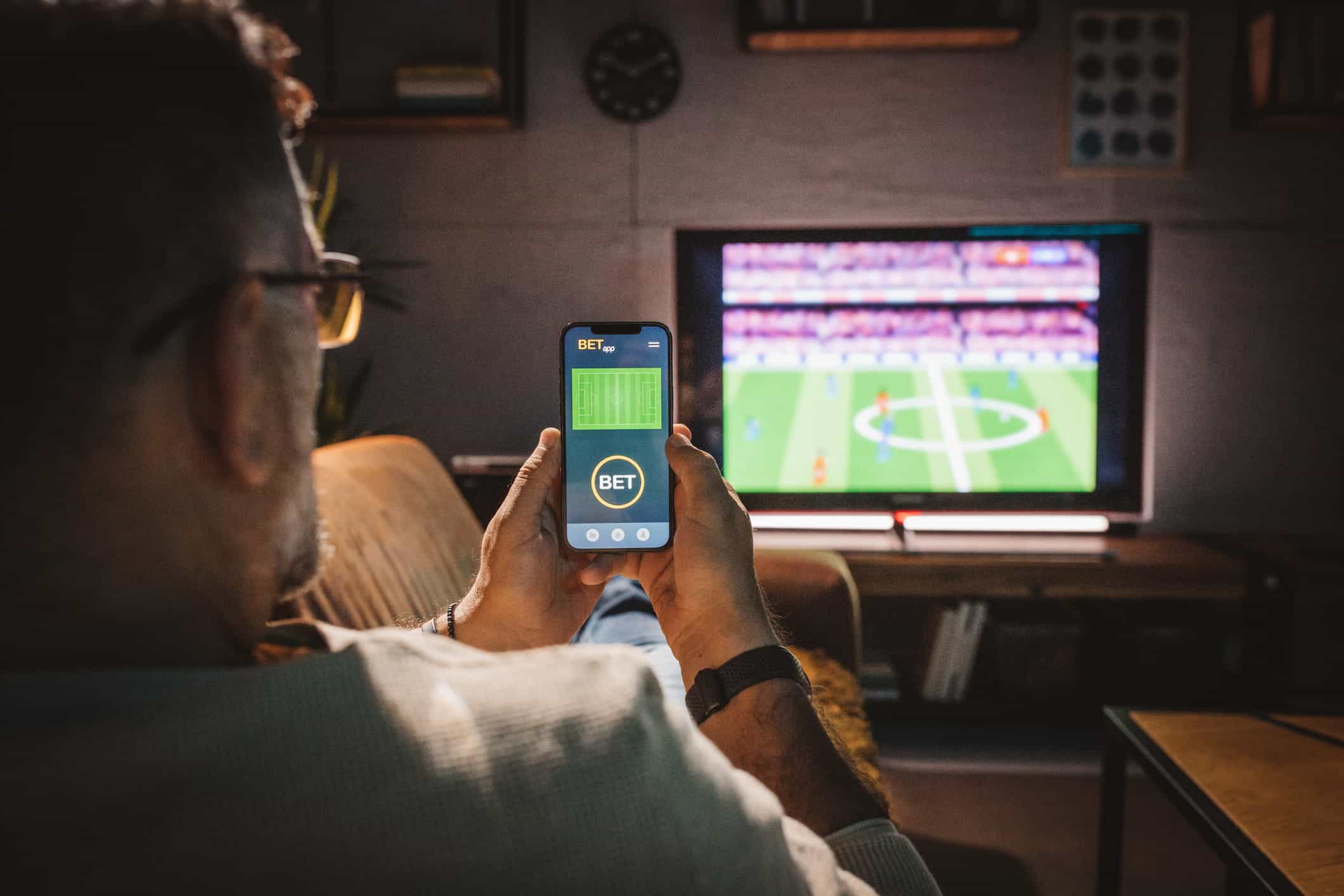 A man sits on a sofa watching a soccer game and using a smart phone to place a bet.