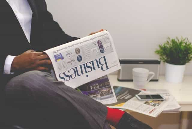 A man in a suit sitting with his legs crossed with a financial newspaper draped over one leg.